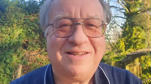 A man head-and-shoulders photo of a man with grey hair and glasses, wearing a blue polo shirt and smiling at the camera. He's outside on a sunny day with trees and a blue sky behind him