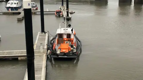 A dredger called Innovation docked at Watchet Marina. The water is a grey/brown colour and wooden walking pontoons are visible