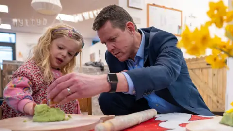 Getty Images Leader of Plaid Cymru Rhun ap Iorwerth during a visit to a nursery. He is sat next to a young girl who has blonde curly hair and is wearing pink. Ap Iorwerth and the girl are playing with green Playdough which is on a wooden board, next to a rolling pin.