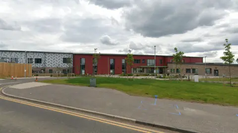 A two-storey building is set back from the road, beyond a grassy area with newly planted trees. The left of Maybury Primary school has a white and grey fascia, with a dark red fascia for the rest of the building and a single storey brick-built wing to the right.