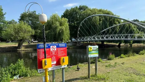 Bedford Borough Council A river in Bedford, showing a butterfly style bridge, with two signs and an emergency board. There is grass on the bank and people walking on the other side of the river. There are a large amount of trees on the other side of the river. 