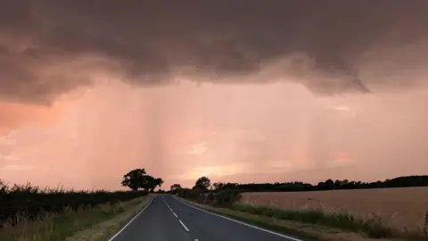 A single carriageway country road with fields on either side. There is a large, dark storm cloud covering the top of the image and there is a pinkish light being cast by the sun which is hidden. There are trees in the distance which look black due to the shadow.