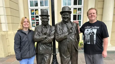 Rebecca Sills - a young woman with blonde hair, wearing a waterproof coat, and Dean Sills - an older man wearing a black Laurel and Hardy T-shirt. They are standing next to statues of Laurel and Hardy. 