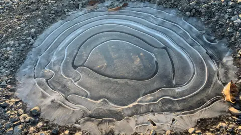 BBC Weather Watchers/Windermere Houlie A frozen puddle on gravel. The ice has formed in uneven, but ever widening concentric circles. Some of the lines are whiter than the surrounding ice, some darker.