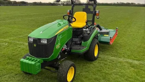 Tregorrick Youth Football Club A picture of a green husqvarna ride-on mower. The mower is on what looks like a football pitch and there are trees and bushes in the background.