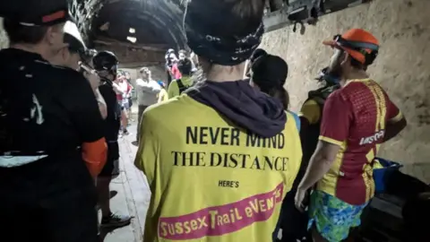 Runners standing in a dimly-lit tunnel ahead of the race. They have their backs to the camera. The person nearest the camera has a yellow jersey printed in the style of the Sex Pistols album cover. It reads: Never Mind the Distance, here's Sussex Trail Events.