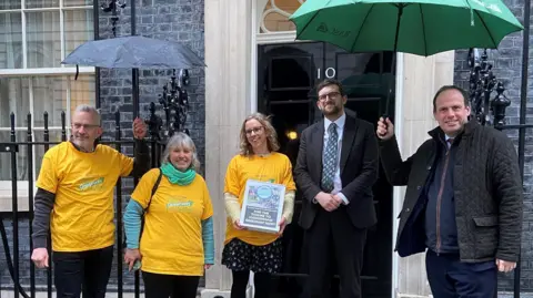 Three campaigners in yellow shirts are joined by MPs Freddie van Mierlo and Greg Smith outside the iconic black door of 10 Downing Street.