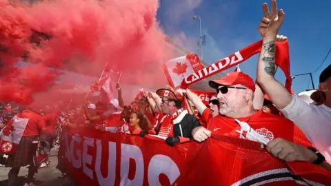 Reuters Fans marching and holding up a big banner that reads 'Voyaguers' in large white letters. They are waving Canadian flags and holding up red scarves that say CANADA in red. Red smoke flairs are blanketing fans in the left half of the photo. 