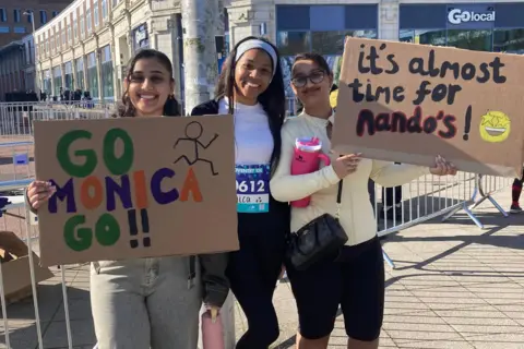 Monica Sahota (pictured centre) is flanked by Anjali Nandra on her right. She is olding a sign reading "Go Monica Go" and on her left Nisha Nandra has a sign saying "I't almost time for Nando's" 