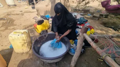 A woman sits outside on the bare ground, washing clothes in a plastic tub. She is wearing a black headscarf that covers much of her face. 