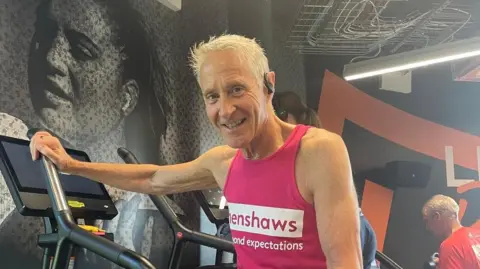 A man with short white hair stands on a stair master. He is wearing a pink vest with white writing