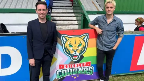 The image shows two men at the edge of a rugby league ground near a white stairway leading up to seating. They are standing on either side of a large sign board displaying the Keighley Cougars club crest, which features a stylised cougar head above the words: KEIGHLEY COUGARS.