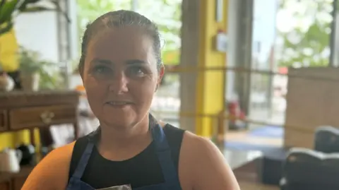 Tess is wearing a blue apron with a grey and white badge on it with her name on it. She has black hair pulled back and is smiling at the camera with plants on her left and the door on her right.