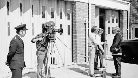 East Sussex Fire and Rescue Service A young Mary Joy Langdon being interviewed by a journalist and camera crew outside a fire station in a black and white image. A male fire officer watches on.