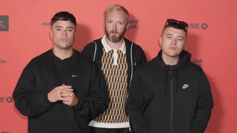 Kneecap members Móglaí Bap, DJ Próvaí and Mo Chara wearing dark clothing and standing against a red background with black writing on it.