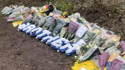 Rows of bouquets of colourful flowers laid on a bank on a dirt path. A football shirt has also been left, as have flowers arranged to spell the word "brother".