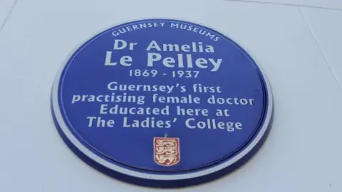 A blue ceramic plaque, whith a gold and red crest at the bottom, hangs on a white wall, on it in white writing reads "Guernsey Museums: Dr Amelia Le Pelley: 1869 - 1937: Guernsey's first practising female doctor Educated here at The Ladies' College."