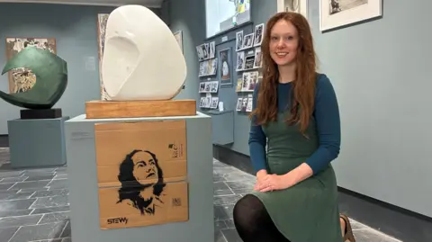 A woman with long red hair smiles at the camera. Next to her is a plinth with a white Barbara Hepworth sculpture. On the plinth is one side of a cardboard box with a stencilled picture of Barbara Hepworth.