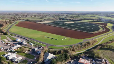 Castle Combe Circuit A drone shot of the circuit - tarmac circuit with bends goes around an area which has grass, a ploughed field area and large numbers of solar panels. Buildings to the left with cars parked. A sunny day.