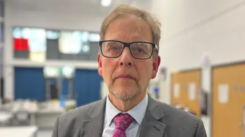 Head and shoulders shot of Tim Boyes, the headteacher. He wears a grey jacket over a blue shirt and pink tie. He has black-rimmed glasses on and blonde hair with a short grey beard.