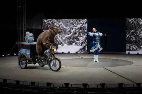 Pietro Rojas A stage performance featuring a large brown bear riding a small three-wheeled motorbike with two colourful nesting-doll props on the back, while a performer in a blue and white costume gestures toward them. A winter forest scene is projected on the screens behind them.
