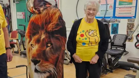 Julianne Moroz A woman in a fancy dress outift that has a lion on it standing in between two people in yellow t-shirts. They are standing in a gym in front of weight machines.