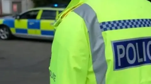 The back of a yellow hi-vis police officer's jacket on the right. It has the words 'POLICE' on the back. On the left, out of focus, is a blue and yellow police car.