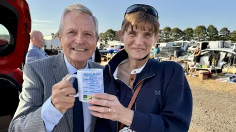 Peter Levy standing next to a woman in a blue jacket. They are holding a mug advertising a dog show. Both are smiling. There are market stalls in the background.
