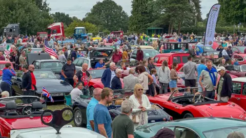 Rows of cars and vehicles, with people gathered around looking at them 