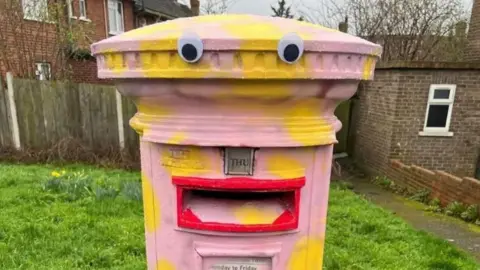 Nik M. A postbox is painted pink with yellow spots. Eyes have been stuck on it and the slot for letters has been painted red to resemble lips. 