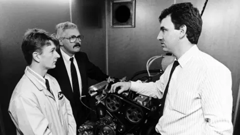 Getty Images A black and white picture of a young Allan McNish in overalls talking to two other men about an engine