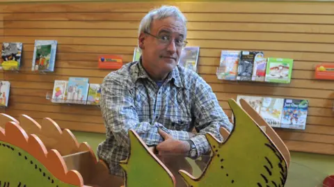 Getty Images Robert Munsch, wearing a checked shirt, poses for the camera with his eyebrows raised. He's leaning on a cut-out of a dragon which appears on the cover of his book the Paper Bag Princess.