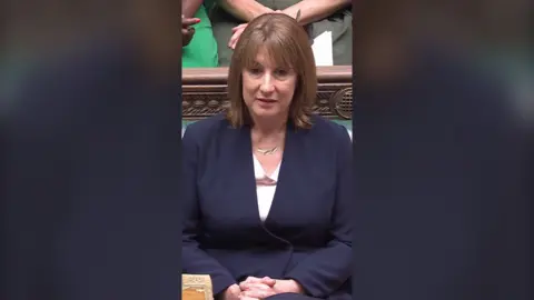 UK Chancellor Rachel Reeves wearing white blouse and navy blazer sitting in the House of Commons bench