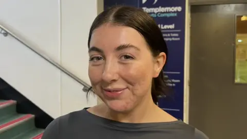 Dearbhla Duffy has long brown hair, which is tied back. She is wearing grey gym gear and standing near a stairwell. 