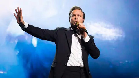Getty Images Olly Murs has brown hair and is singing into a microphone on stage. One arm is out as he sings. He is wearing a black suit jacket, black trousers, a black bow tie and white shirt. The lighting behind him is blue