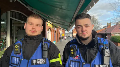 Gus and Callum looking into the camera, stood next to each other on one of Oswestry's shopping streets. They're wearing royal blue vests with radios and body-worn cameras, similar to what police wear. They're stood underneath a dark green awning, and you can see the street stretching away behind them.