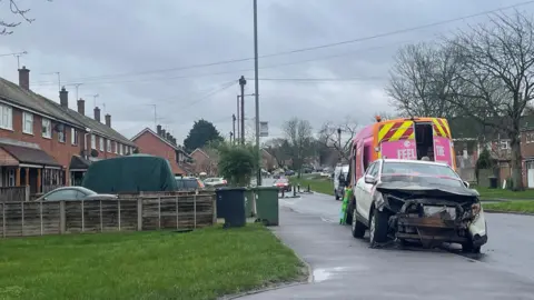 A white car, which has been burnt out, is parked on the pavement in this picture taken on a road in Warndon West. Along the left hand side is a row of houses and gardens,