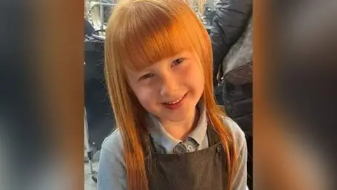 Lila with ginger hair and grey pinafore and light grey top smiling.