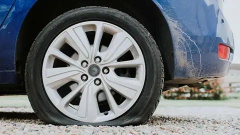 A close up of a blue car with silver alloys and a flat tyre. Dried mud covers the front bumper of the car, which is parked on white gravel.