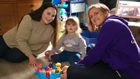 Martin Giles/BBC Heather and Freya with a home-start volunteer sitting on the floor playing with toys. The mother has long reddish dark hair and is wearing ribbed beige sweater. The female volunteer has shoulder length blonde hair with red streaks and is wearing a a purple hoodie. The toddler is wearing blue jeans and a brown sweatshirt.
