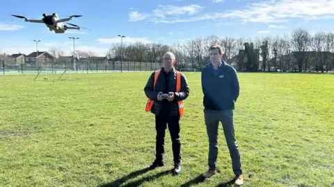 Two council employees watch as one of them flies a drone a few feet off the ground on a playing field