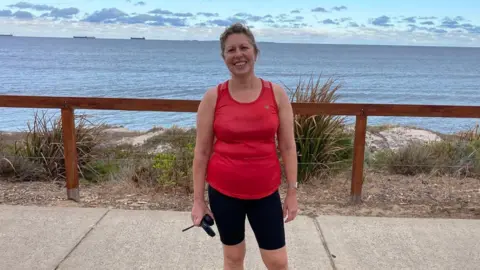 Elaine Wan out running. She is wearing a red vest top and black shorts. She is smiling and there is a beach behind her. 