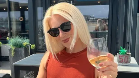 Facebook A woman with sunglasses on and long blonde hair looks at the camera. She has a burnt orange dress on and holds up a glass of white wine. It is a head and shoulders shot. 