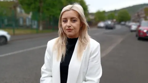A woman with long blonde hair wearing a cream jacket and black top stands on the footpath beside a road. Cars and houses can be seen in the background.