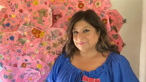 A smiling woman stands in front of a pink quilt made up of hexagonal patches with flowers on them