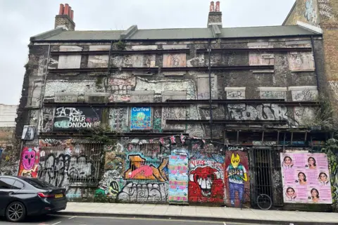 Three brick buildings covered in graffiti with windows boarded up and metal fences across the front