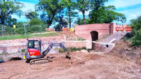 Wilts and Berks Canal Trust A digger is clearing a large area of earth in the foreground and behind it there is a section of brick wall and a bridge with a narrow entrance through it