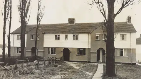 Express & Star/Wolverhampton City Archives. A black and white photo of terraced homes on two storeys. The homes are white at the top with brickwork on the bottom storey. They also have chimneys. In front of the homes are several trees and some grass areas with a wooden fence. 