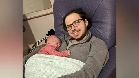 Family handout A man smiles while sat in a chair holding a newborn baby. The man has glasses and long dark hair. They are both covered in a blue blanket. 