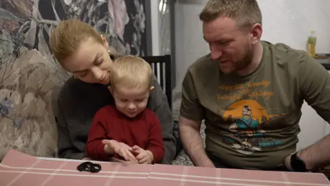 A little boy is sitting on a woman's lap at a table, his father beside him, and a toy car on the table